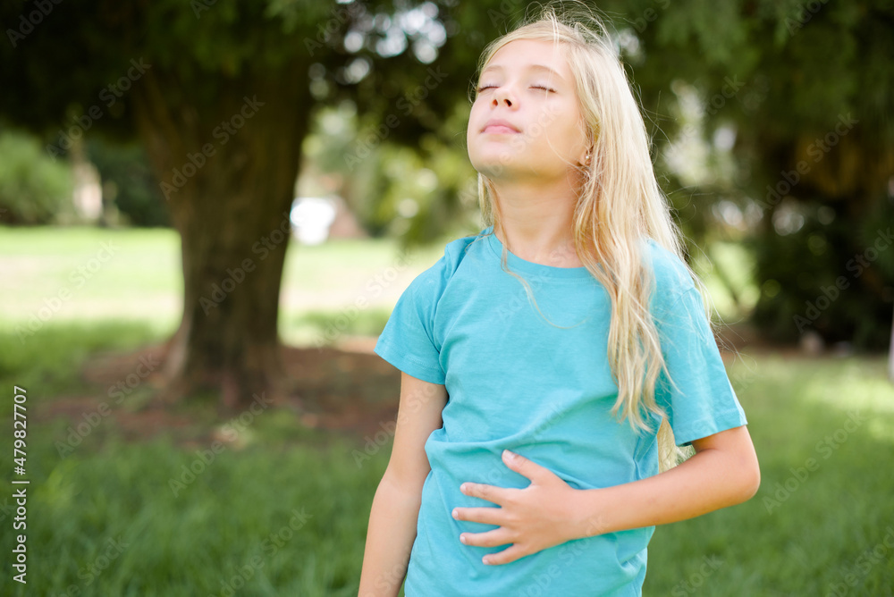 Caucasian little kid girl wearing blue T-shirt standing outdoors touches tummy, smiles gently, eating and satisfaction concept. Caucasian little kid girl wearing blue T-shirt standing outdoors touches tummy, smiles gently, eating and satisfaction concept.