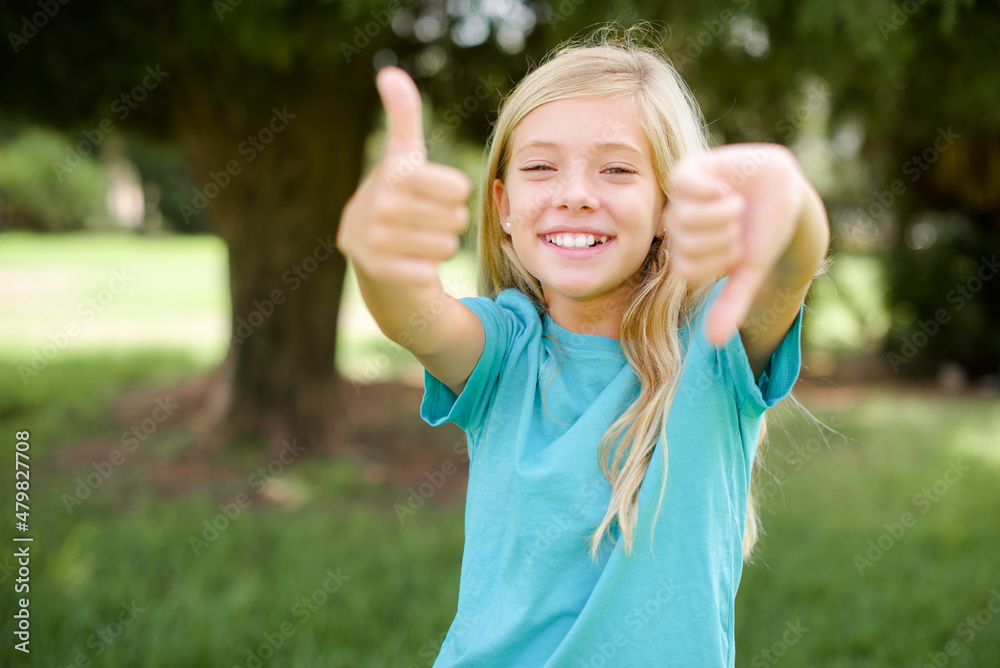 Caucasian little kid girl wearing blue T-shirt standing outdoors showing thumbs up and thumbs down, difficult choose concept Caucasian little kid girl wearing blue T-shirt standing outdoors showing thumbs up and thumbs down, difficult choose concept