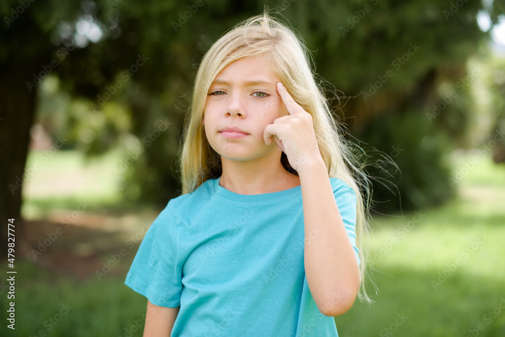 Caucasian little kid girl wearing blue T-shirt standing outdoors tries to memorize something, keeps fore finger on temple, reminds information for exam,