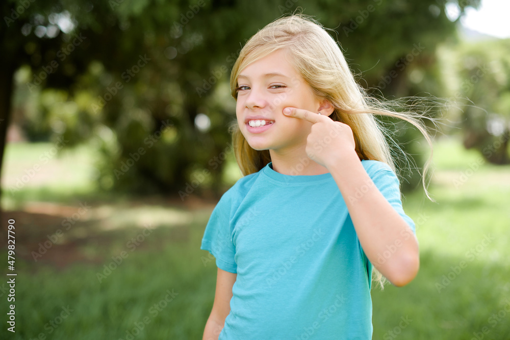 Caucasian little kid girl wearing blue T-shirt standing outdoors pointing unhappy to pimple on forehead, ugly infection of blackhead. Acne and skin problem