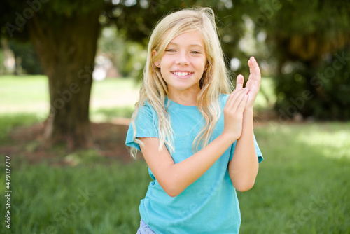 Caucasian little kid girl wearing blue T-shirt standing outdoors clapping and applauding happy and joyful, smiling proud hands together.