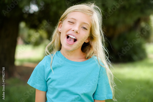 Canvas Print Caucasian little kid girl wearing blue T-shirt standing outdoors winking looking at the camera with sexy expression, cheerful and happy face