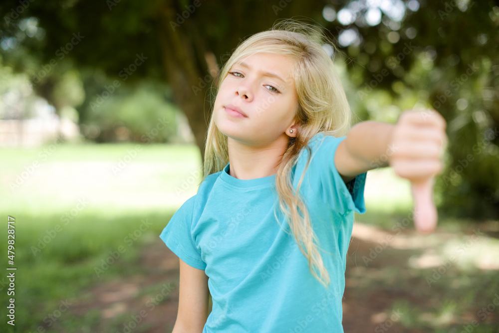 Caucasian little kid girl wearing blue T-shirt standing outdoors feeling angry, annoyed, disappointed or displeased, showing thumbs down with a serious look Caucasian little kid girl wearing blue T-shirt standing outdoors feeling angry, annoyed, disappointed or displeased, showing thumbs down with a serious look