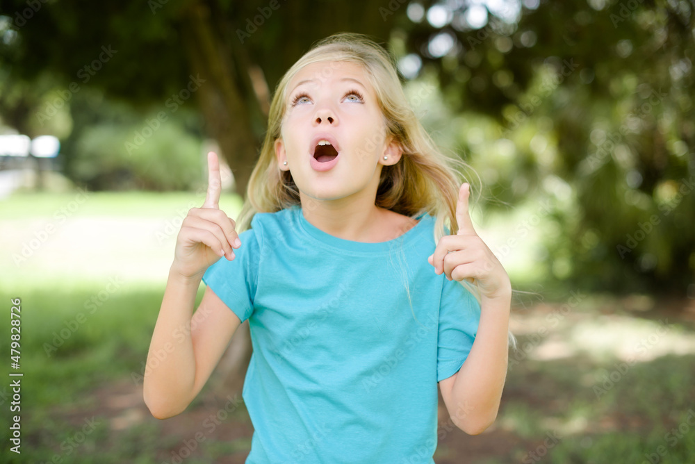 Caucasian little kid girl wearing blue T-shirt standing outdoors amazed and surprised looking up and pointing with fingers and raised arms. Caucasian little kid girl wearing blue T-shirt standing outdoors amazed and surprised looking up and pointing with fingers and raised arms.