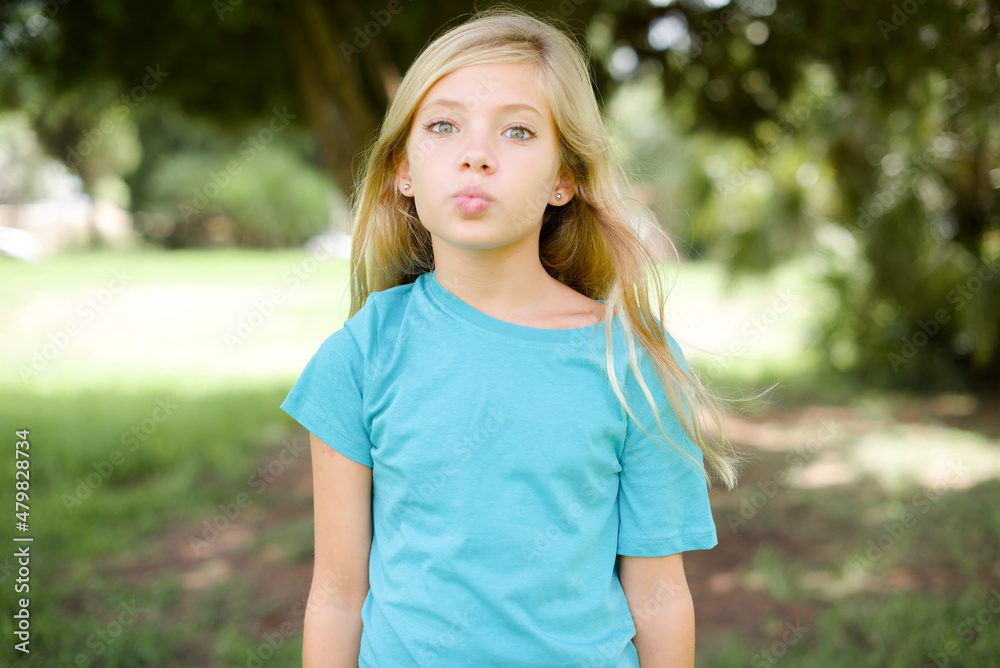 Shot of pleasant looking Caucasian little kid girl wearing blue T-shirt standing outdoors , pouts lips, looks at camera, Human facial expressions Shot of pleasant looking Caucasian little kid girl wearing blue T-shirt standing outdoors , pouts lips, looks at camera, Human facial expressions