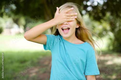 Caucasian little kid girl wearing blue T-shirt standing outdoors smiling and laughing with hand on face covering eyes for surprise. Blind concept.