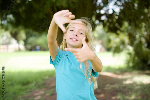 Caucasian little kid girl wearing blue T-shirt standing outdoors making finger frame with hands. Creativity and photography concept.