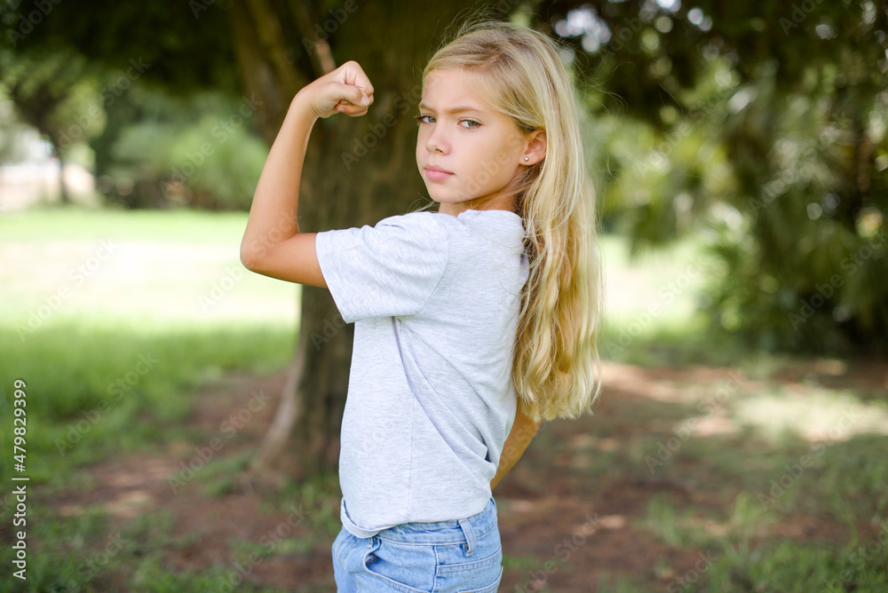 Portrait of powerful cheerful Caucasian little kid girl wearing white T-shirt standing outdoors showing muscles.