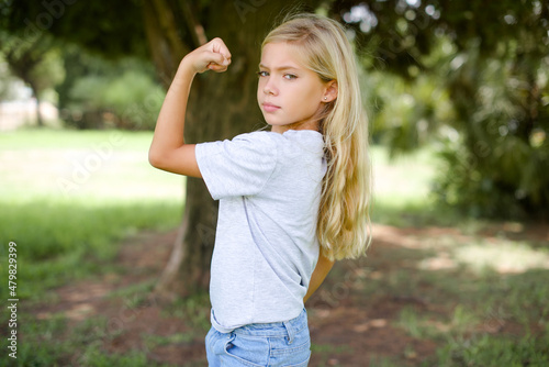 Portrait of powerful cheerful Caucasian little kid girl wearing white T-shirt standing outdoors showing muscles.