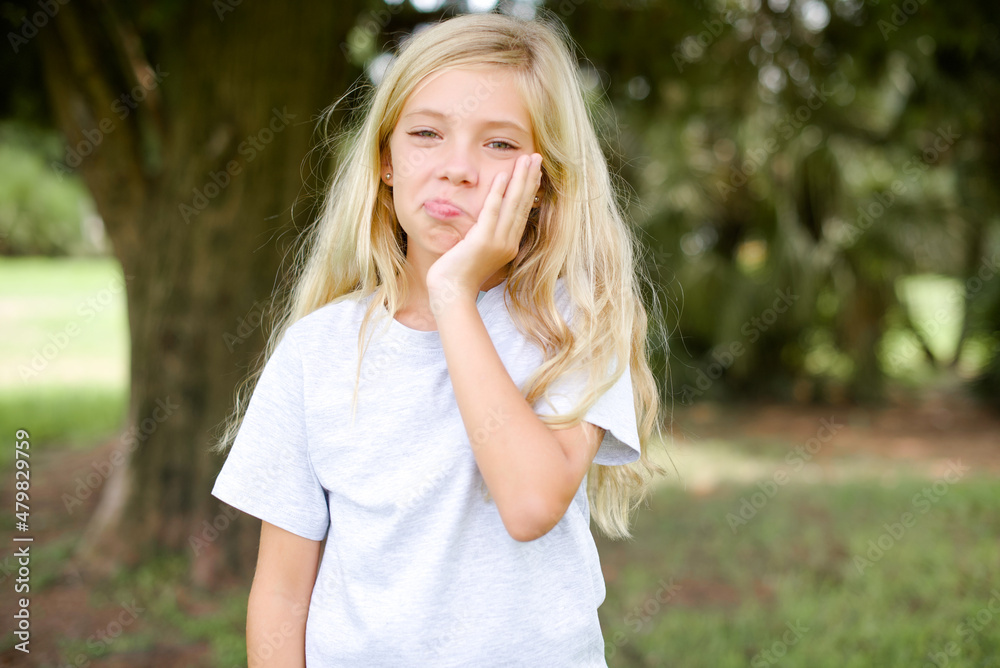 Sad lonely Caucasian little kid girl wearing whiteT-shirt standing outdoors touches cheek with hand bites lower lip and gazes with displeasure. Bad emotions