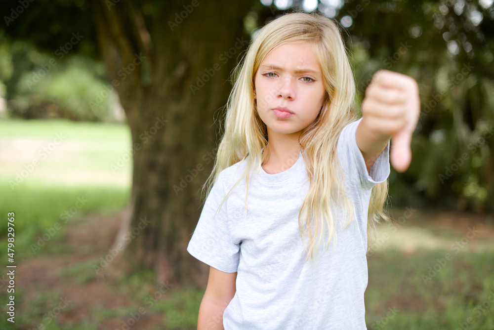 Caucasian little kid girl wearing whiteT-shirt standing outdoors looking unhappy and angry showing rejection and negative with thumbs down gesture. Bad expression. Caucasian little kid girl wearing whiteT-shirt standing outdoors looking unhappy and angry showing rejection and negative with thumbs down gesture. Bad expression.