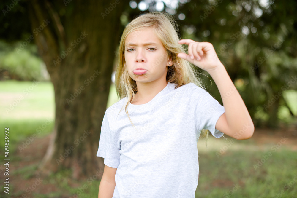 Caucasian little kid girl wearing whiteT-shirt standing outdoors purses lip and gestures with hand, shows something very little. Caucasian little kid girl wearing whiteT-shirt standing outdoors purses lip and gestures with hand, shows something very little.