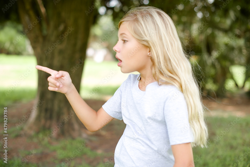Stunned beautiful Caucasian little kid girl wearing whiteT-shirt standing outdoors with greatly surprised expression points away on copy space, indicates something Stunned beautiful Caucasian little kid girl wearing whiteT-shirt standing outdoors with greatly surprised expression points away on copy space, indicates something