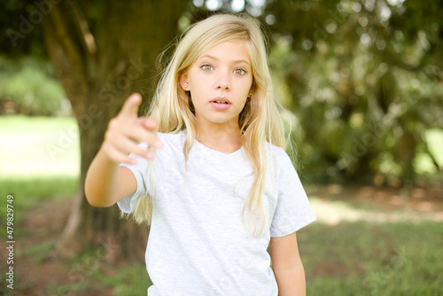 Fototapeta Naklejka Na Ścianę i Meble -  Shocked Caucasian little kid girl wearing whiteT-shirt standing outdoors points at you with stunned expression