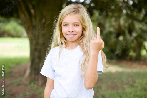Caucasian little kid girl wearing whiteT-shirt standing outdoors shows middle finger bad sign asks not to bother. Provocation and rude attitude. photo