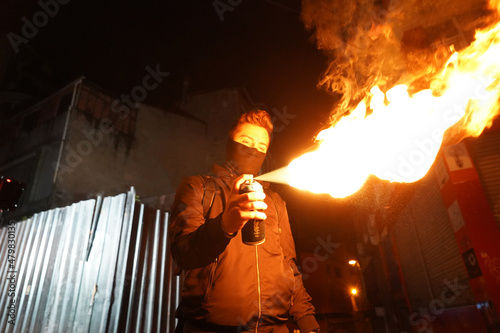 A young graffiti artist in a black hoodie looks at the wall Street art concept, Vandal boy with hidden face makes fire from spray can gases.
