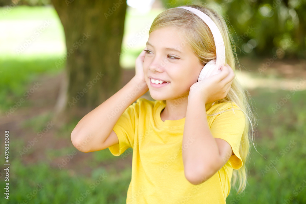 beautiful Caucasian little kid girl wearing yellow T-shirt standing outdoors wears stereo headphones listens music concentrated aside. People hobby lifestyle concept