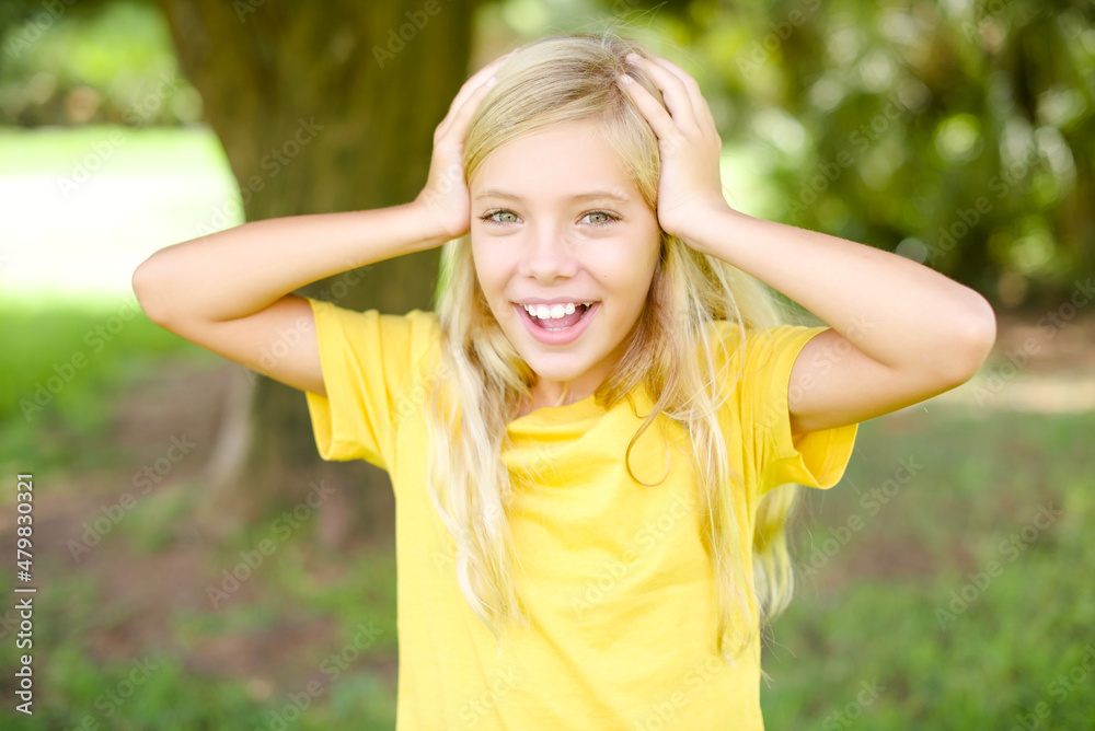 Cheerful overjoyed beautiful Caucasian little kid girl wearing yellow T-shirt standing outdoors reacts rising hands over head after receiving great news. Cheerful overjoyed beautiful Caucasian little kid girl wearing yellow T-shirt standing outdoors reacts rising hands over head after receiving great news.