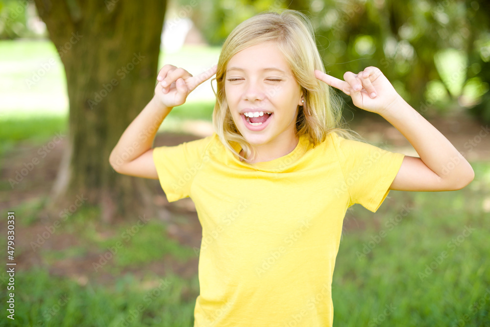 Photo of crazy beautiful Caucasian little kid girl wearing yellow T-shirt standing outdoors screaming and pointing with fingers at hair closed eyes Photo of crazy beautiful Caucasian little kid girl wearing yellow T-shirt standing outdoors screaming and pointing with fingers at hair closed eyes