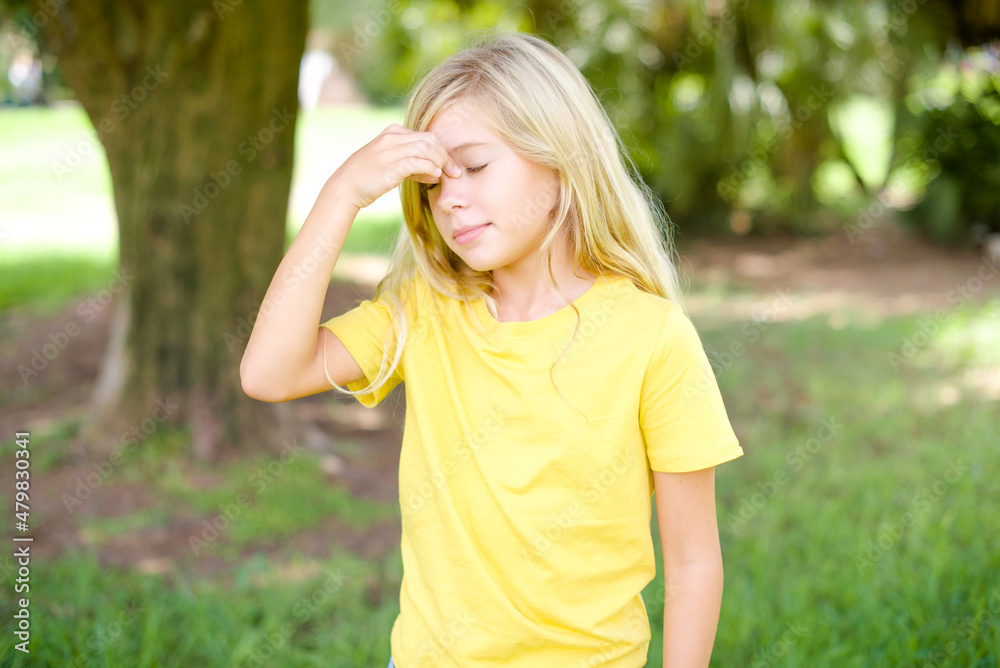 Sad beautiful Caucasian little kid girl wearing yellow T-shirt standing outdoors suffering from headache holding hand on her face Sad beautiful Caucasian little kid girl wearing yellow T-shirt standing outdoors suffering from headache holding hand on her face