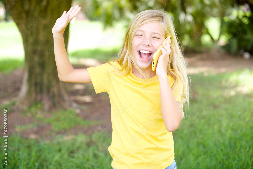 Overemotive happy beautiful Caucasian little kid girl wearing yellow T-shirt standing outdoors laughs out positively hears funny story from friend during telephone conversation