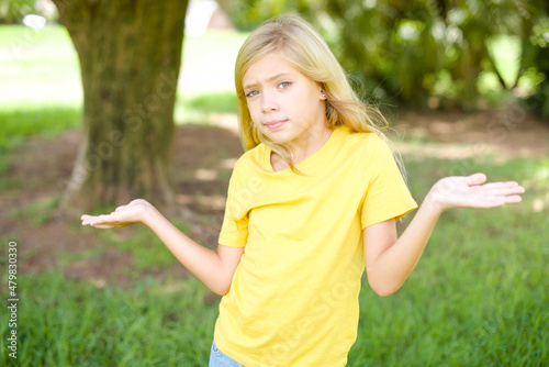 Careless attractive beautiful Caucasian little kid girl wearing yellow T-shirt standing outdoors shrugging shoulders, oops.