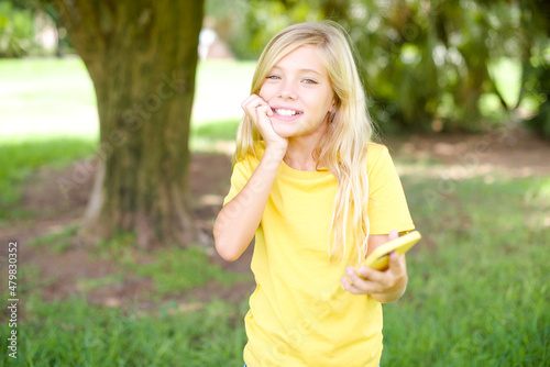 Afraid funny beautiful Caucasian little kid girl wearing yellow T-shirt standing outdoors holding telephone and bitting nails