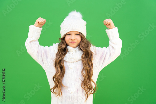 A girl in a warm white sweater and hat shows her fist and is very angry. A disgruntled baby with curly hair on an isolated background.