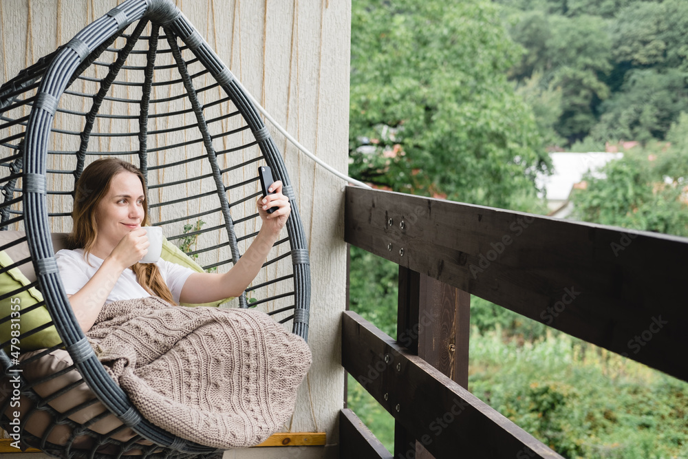 Young woman sitting in a hanging chair with a blanket and a cup of coffee on the balcony.