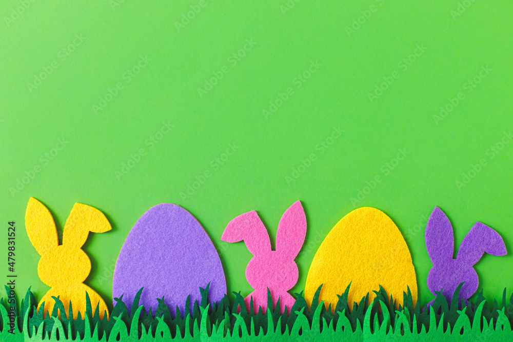 Easter hunt concept. Colorful Easter bunnies and eggs in grass on green background, top view with space for text. Happy Easter! Pink, yellow, purple artificial bunny and egg decor.