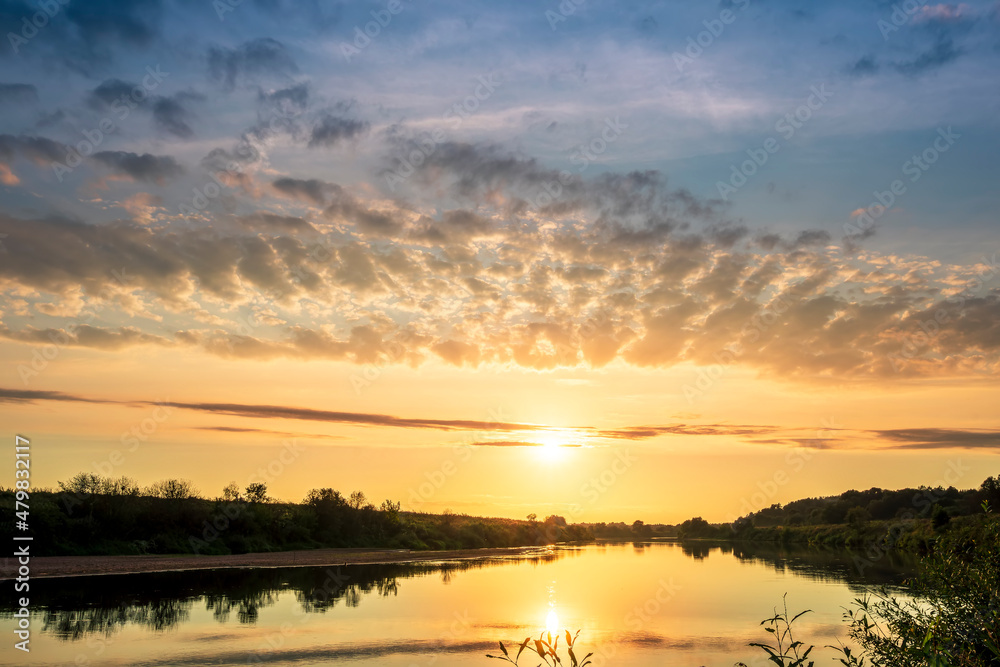 Fototapeta premium Scenic view at beautiful sunset or sunrise on a shiny river with green bushes on sides, golden sun rays, calm water ,deep blue cloudy sky and forest on a background, spring landscape