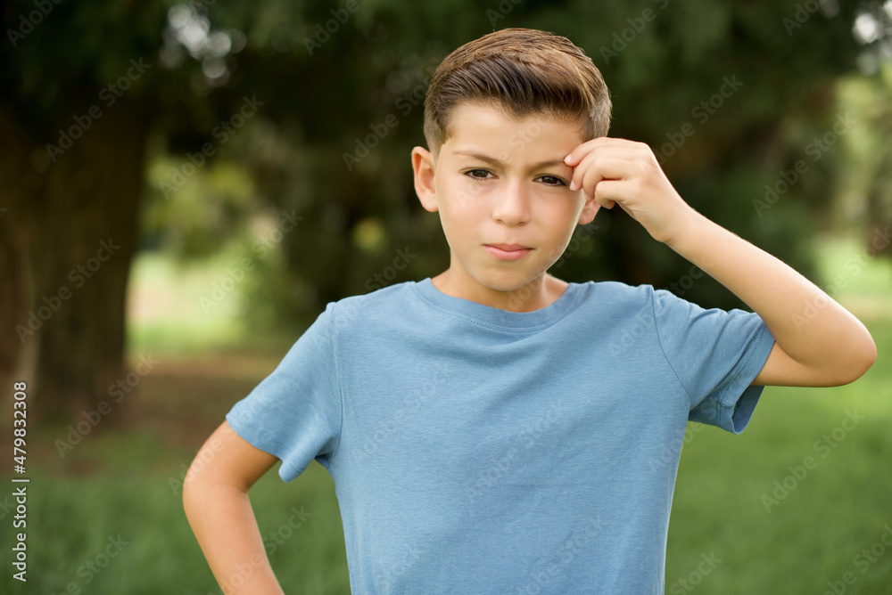 Caucasian little kid boy wearing blue T-shirt standing outdoor pointing unhappy at pimple on forehead, blackhead infection. Skincare concept. Caucasian little kid boy wearing blue T-shirt standing outdoor pointing unhappy at pimple on forehead, blackhead infection. Skincare concept.