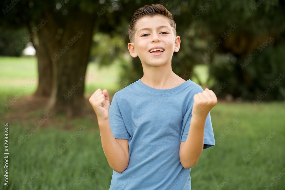 Caucasian little kid boy wearing blue T-shirt standing outdoor celebrating surprised and amazed for success with arms raised and eyes closed. Winner concept. Caucasian little kid boy wearing blue T-shirt standing outdoor celebrating surprised and amazed for success with arms raised and eyes closed. Winner concept.