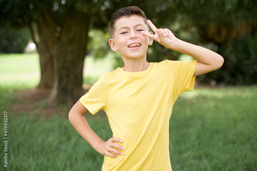 Caucasian little kid boy wearing yellow T-shirt standing outdoor Doing peace symbol with fingers over face, smiling cheerful showing victory Caucasian little kid boy wearing yellow T-shirt standing outdoor Doing peace symbol with fingers over face, smiling cheerful showing victory
