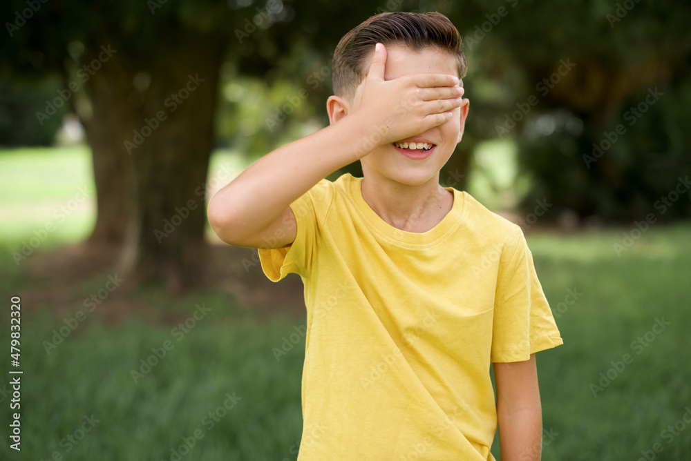 Caucasian little kid boy wearing yellow T-shirt standing outdoor smiling and laughing with hand on face covering eyes for surprise. Blind concept. Caucasian little kid boy wearing yellow T-shirt standing outdoor smiling and laughing with hand on face covering eyes for surprise. Blind concept.
