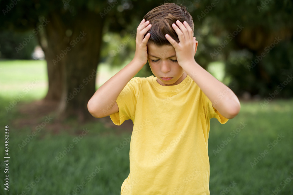 Caucasian little kid boy wearing yellow T-shirt standing outdoor  suffering from strong headache desperate and stressed because of overwork. Depression and pain concept.