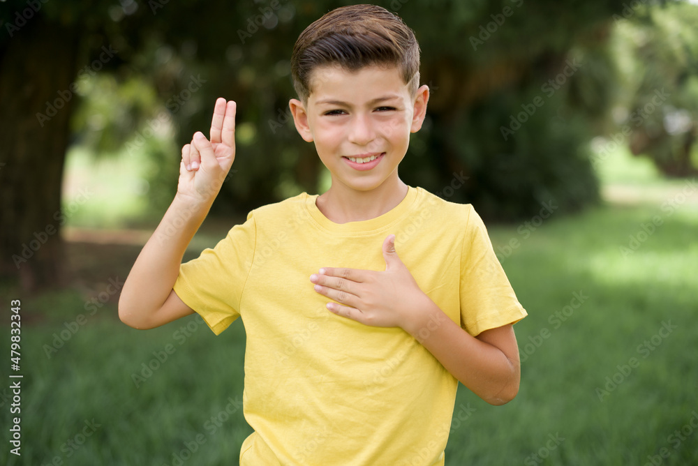 Caucasian little kid boy wearing yellow T-shirt standing outdoor smiling swearing with hand on chest and fingers up, making a loyalty promise oath. Caucasian little kid boy wearing yellow T-shirt standing outdoor smiling swearing with hand on chest and fingers up, making a loyalty promise oath.