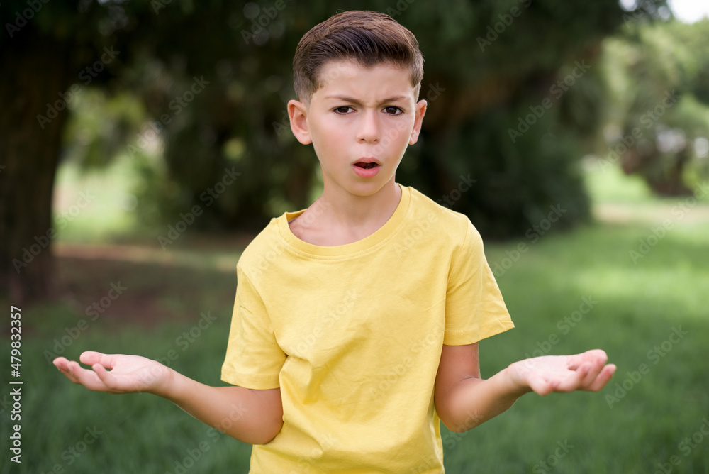Frustrated Caucasian little kid boy wearing yellow T-shirt standing outdoor feels puzzled and hesitant, shrugs shoulders in bewilderment, keeps mouth widely opened, doesn't know what to do. Frustrated Caucasian little kid boy wearing yellow T-shirt standing outdoor feels puzzled and hesitant, shrugs shoulders in bewilderment, keeps mouth widely opened, doesn't know what to do.