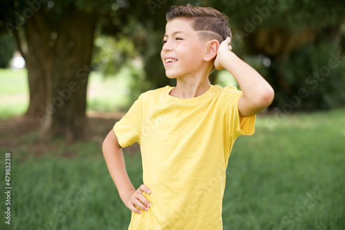 Caucasian little kid boy wearing yellow T-shirt standing outdoor confuse and wonder about question. Uncertain with doubt, thinking with hand on head. Pensive concept.
