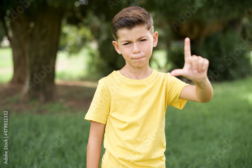 Caucasian little kid boy wearing yellow T-shirt standing outdoor making fun of people with fingers on forehead doing loser gesture mocking and insulting.