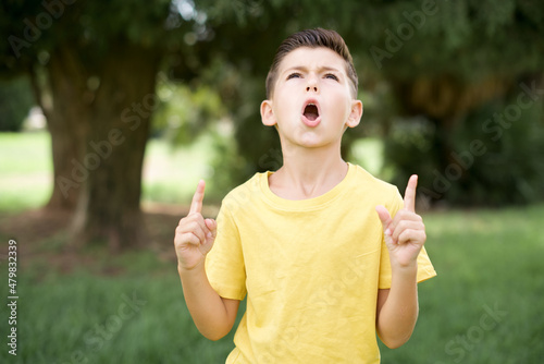 Caucasian little kid boy wearing yellow T-shirt standing outdoor  amazed and surprised looking up and pointing with fingers and raised arms.