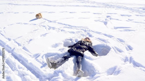 Young little girl in warm jacket with fur, jeans, boots and gloves lying on white clean snow doing snow angel, playing, having fun with her dog pet Pomeranian Spitz on sunny winter day. Holidays