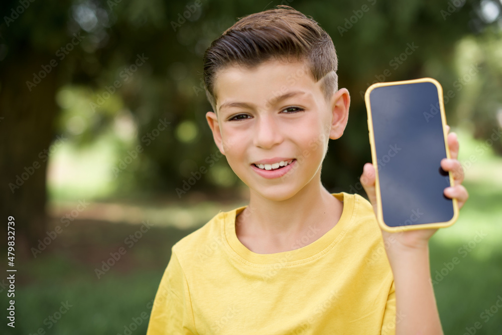 Smiling Caucasian little kid boy wearing yellow T-shirt standing outdoors Mock up copy space. Hold mobile phone with blank empty screen Smiling Caucasian little kid boy wearing yellow T-shirt standing outdoors Mock up copy space. Hold mobile phone with blank empty screen