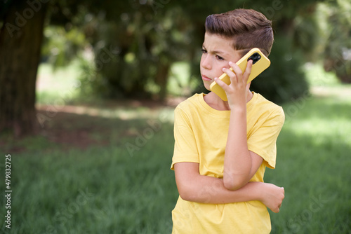 Sad Caucasian little kid boy wearing yellow T-shirt standing outdoors  talking on smartphone. Communication concept.