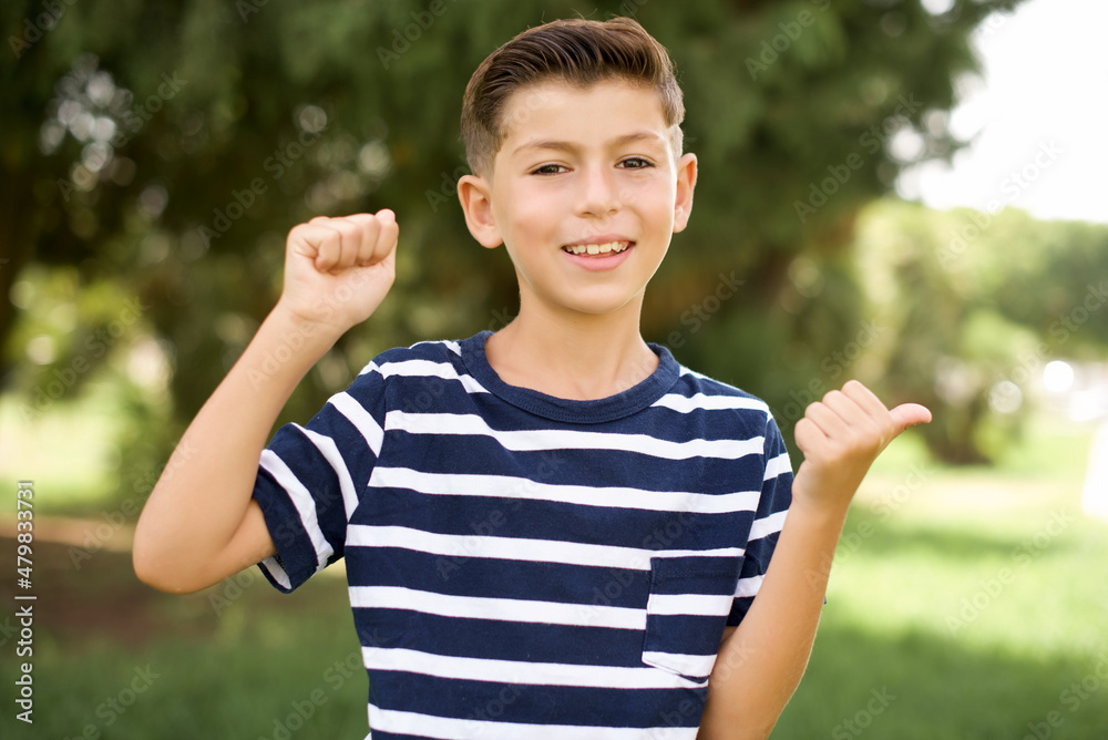 Hooray cool beautiful Caucasian little kid boy wearing stripped T-shirt standing outdoors point back empty space hand fist Hooray cool beautiful Caucasian little kid boy wearing stripped T-shirt standing outdoors point back empty space hand fist