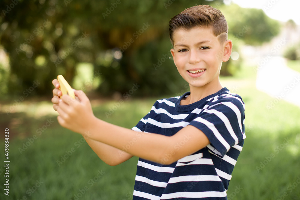 Nice addicted cheerful beautiful Caucasian little kid boy wearing stripped T-shirt standing outdoors using gadget playing network game Nice addicted cheerful beautiful Caucasian little kid boy wearing stripped T-shirt standing outdoors using gadget playing network game