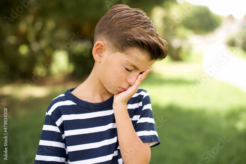 beautiful Caucasian little kid boy wearing stripped T-shirt standing outdoors with sad expression covering face with hands while crying. Depression concept.