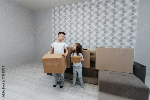 Brothers and sister are playing and enjoying moving to a new apartment against the background of cardboard boxes and a sofa. Purchase of real estate. Housewarming, delivery. © Andrii