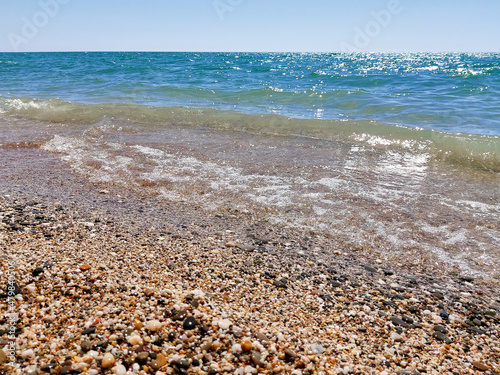 Sea pebbles, blue sea, waves, bright sun. The bright blue surface of the sea. Landscape wallpaper and nature concept. Warm summer sea with small waves in good weather.