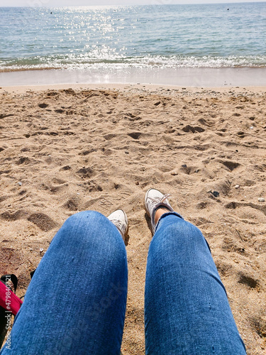 A woman is sitting on the seashore. Woman legs in white sneakers and jeans on yellow sand beach in summer day. Ocean clear water seafoam, yellow sand, small wawes. Summer background.